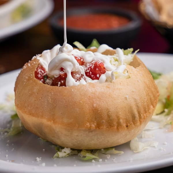 A Mexican tostada bowl filled with diced tomatoes, lettuce, and sour cream being drizzled.