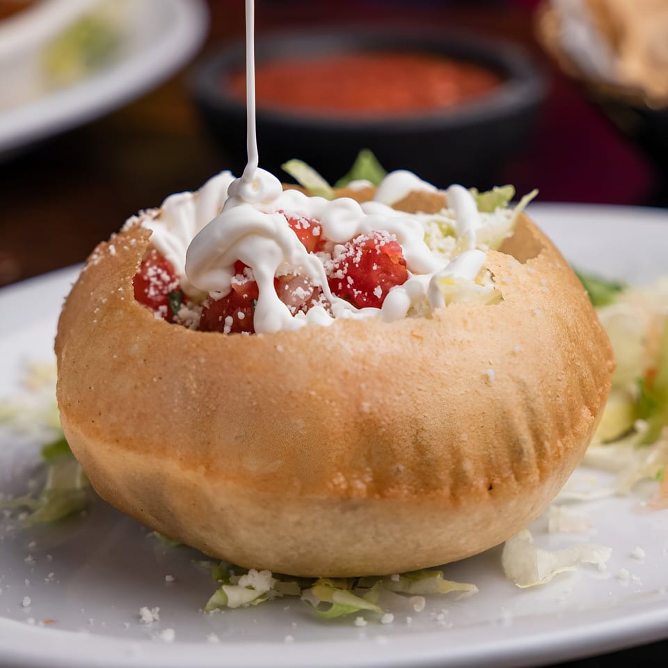 A Mexican tostada bowl filled with diced tomatoes, lettuce, and sour cream being drizzled.